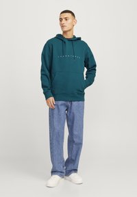 Teal hettegenser med frontlomme, snor og logo trykk; kombinert med løse blå jeans og hvite sneakers.