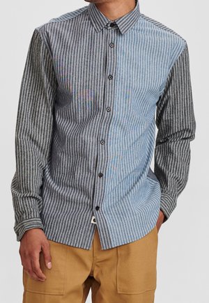 Homme portant une chemise boutonnée à manches longues avec des rayures verticales noires, grises et bleues et un pantalon beige, mains détendues le long du corps.