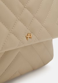 Sac en cuir matelassé beige avec une couture diagonale et un petit logo en métal doré sur le rabat avant.