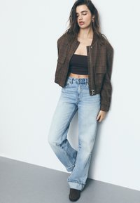 Ruskean ruudullinen bomber-takki, jossa on lyhennetty muotoilu, yhdistetty kevyisiin sinisiin levennettyihin farkkuihin. Mallilla on yllä tummanruskea crop-top ja ruskeat kengät.