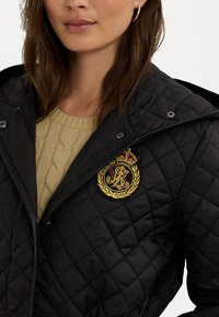 Svart vadderad jacka med huva, med en framträdande broderad emblem med en krona och lagerdesign. Bärs över en beige kabelstickad tröja.