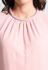 Blouse rose clair avec un col froncé et une bordure texturée ornée de perles. Tissu lisse, coupe ample et détails de plis subtils.