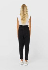 Donna con lunghe onde bionde, che indossa un top bianco senza maniche, pantaloni neri a vita alta e sneakers bianche, in piedi di spalle.