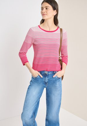 Kobieta stojąca z rękami w kieszeniach, ubrana w czerwono-białą koszulę w paski z długim rękawem, niebieskie dżinsy oraz brązową torbę na ramię.