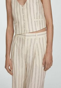 Vrouwelijk bovenlijf in een beige mouwloze gewatteerde vest met bijpassende hoog-taille broek met dunne verticale zwarte strepen.