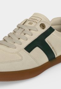 Ανδρικό sneaker Tommy Hilfiger εκρού με κρεμ κορδόνια, σκούρα πράσινη πλαϊνή ρίγα, λαστιχένια σόλα και μεταλλική οπή με λογότυπο και τυπωμένα λογότυπα.