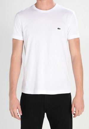 T-shirt blanc en coton à manches courtes, col rond, avec un logo vert brodé sur le côté gauche de la poitrine. Porté avec un pantalon noir.