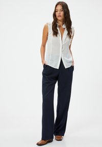 Blusa blanca sin mangas con cuello con volantes, combinada con pantalones de pinstripe azul marino y mocasines marrones. Tejido liso con un ajuste ajustado.