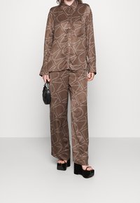 Malli, joka pukeutuu ruskeaan satiinishirtamiseen ja leveiden lahkeiden housuihin, joissa on valkoinen abstrakti viivakuosis, pitelee mustaa tikattua laukkua ja mustia korkokenkiä.