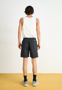 Melnas vieglas šortus ar elastīgu jostasvietu, sānu kabatu un vienkāršu baltu bezpiegu krekliņu, skatīts no aizmugures uz dzeltena grīdas.