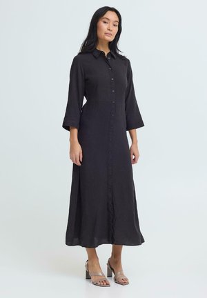 Kobieta w długiej czarnej sukience zapinanej na guziki, z rękawami o długości do łokcia, w beżowych sandałach na obcasie, stojąca na tle jasnego, jednolitego tła.