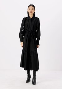 Musta shirt-mekko, jossa on nappikaulus, vyötärölle sidottava vyö ja kerroksellinen helma. Valmistettu teksturoidusta kankaasta, jossa on hienovarainen kuvio. Yhdistetty polven yli ulottuviin saappaisiin.