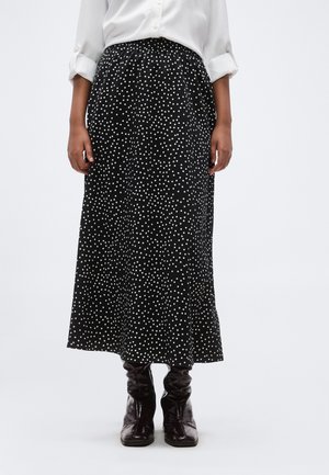 Frau trägt einen schwarzen Midirock mit weißen Punkten, kombiniert mit einem weißen Langarmhemd und dunkelbraunen Lederstiefeln.