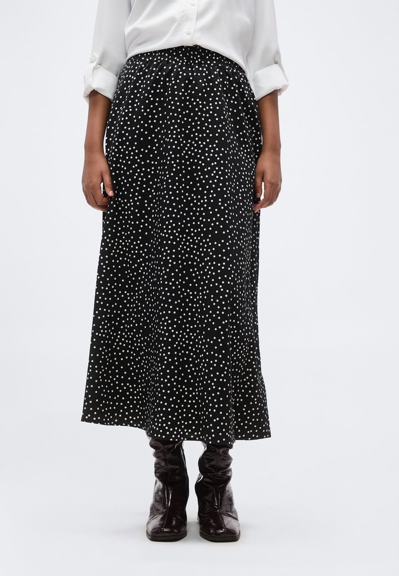 Γυναίκα που φοράει μαύρη μίντι φούστα με λευκές πουά, συνδυασμένη με λευκό πουκάμισο μακρυμάνικο και σκούρα καφέ δερμάτινες μπότες.