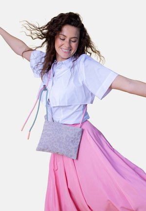 Woman with curly hair smiling and spinning, wearing a white cropped shirt, long pink skirt, and light purple patterned crossbody bag.