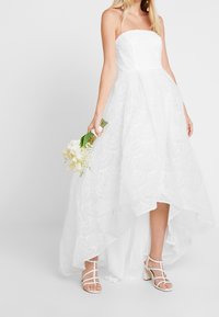 Robe blanche sans bretelles avec un ourlet en high-low, texture en dentelle superposée et motif floral. Assortie avec des sandales blanches à lanières et un bouquet.