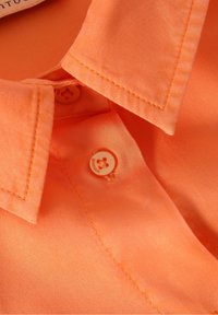 Gros plan sur le col d'une chemise orange vif et deux boutons assortis avec des coutures visibles sur un tissu lisse.