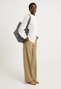 Blusa blanca bordada, pantalones de pierna ancha beige y un bolso de mano oscuro con estampado. La modelo lleva zapatos de punta color marrón claro. Fondo simple.