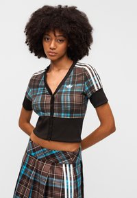 Καφέ και μπλε καρό cropped τοπ με κοντά μανίκια, μαύρες λεπτομέρειες, V-λαιμόκοψη και κούμπωμα με κουμπιά. Συνοδευτική καρό φούστα με πιέτες και ρίγες.