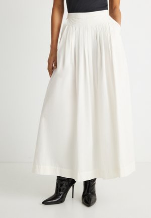 Kvinde iført en hvid plisseret midinederdel med høj talje og sidelommer samt sorte spidse stiletstøvler.