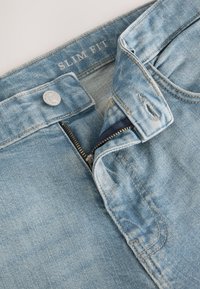 Jean en denim bleu clair, coupe slim, avec fermeture éclair en métal et bouton. Présente des coutures visibles et un tissu doux et texturé.