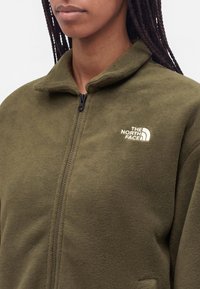 Femme portant une veste polaire zippée vert olive avec le logo The North Face sur le côté gauche de la poitrine, debout devant un fond blanc.