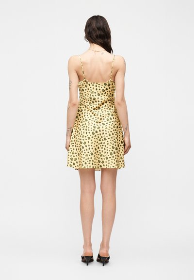 Γυναίκα με μακριά σκούρα μαλλιά που φοράει μανίκι-κίτρινο φόρεμα με σχέδιο λεοπάρδαλης και μαύρα ψηλοτάκουνα σανδάλια, στέκεται με την πλάτη προς τα πίσω σε λευκό φόντο.