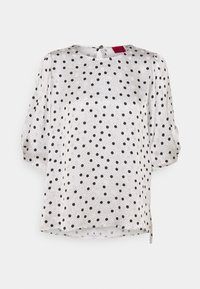 Witte blouse met zwarte stippen en puffed driekwartmouwen, voorzien van een sluiting met knopen aan de achterkant en een ronde halslijn.