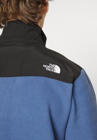 Czarna i niebieska kurtka polarowa z wysokim kołnierzem, z białym logo na ramieniu. Miękka faktura i kontrastujące kolorowe sekcje.