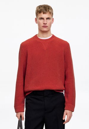 Junger Mann, der einen rostfarbenen Strickpullover und schwarze Hose trägt, vor einem schlichten weißen Hintergrund steht und eine Tasche in der linken Hand hält.