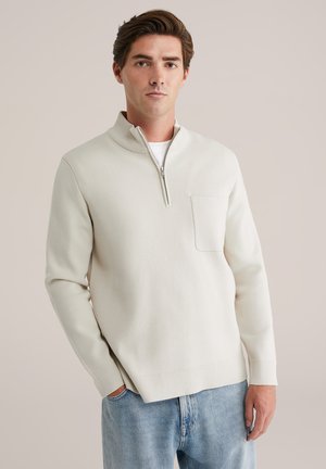 Maglione beige con zip a metà e finitura testurizzata, dotato di tasca sul petto, colletto e polsini a costine, abbinato a jeans blu chiaro.