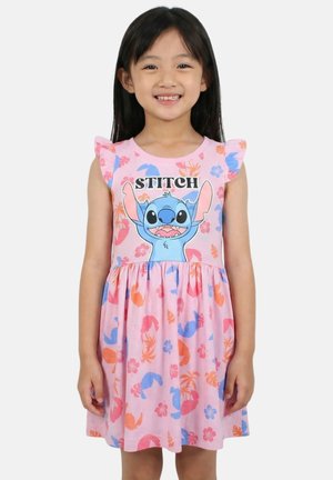 Jeune fille souriante, portant une robe rose avec des manches à volants, avec le personnage bleu Stitch et des motifs floraux et feuillus colorés.