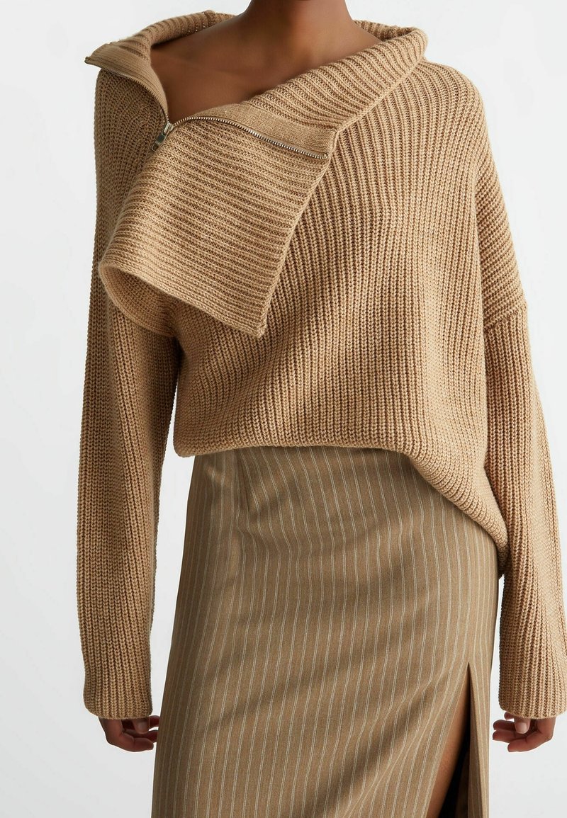 Pull tricoté beige avec un gros maillage et un grand détail de fermeture éclair pliée décalée sur l'épaule, associé à une jupe à fines rayures beige avec une fente à l'avant.