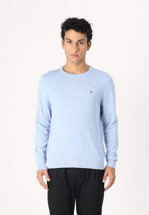 Mann iført lyseblå genser med lange ermer og liten logo på brystet, og svarte bukser, vender fronten mot en ensfarget hvit bakgrunn.