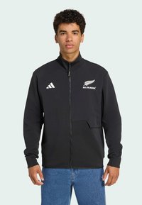 Sort sportjakke med høj krave, fuld lynlås, Adidas-logo, All Blacks-emblem og lomme detaljer. Lavet af glat, strækbart stof.