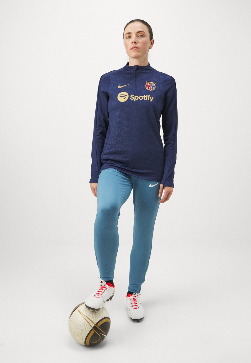 Marineblaues Langarmtrikot mit Viertel-Reißverschluss, ausgestattet mit einem goldenen Spotify-Logo und dem FC Barcelona-Wappen, kombiniert mit hellblauen eng anliegenden Hosen und weißen Fußballschuhen.