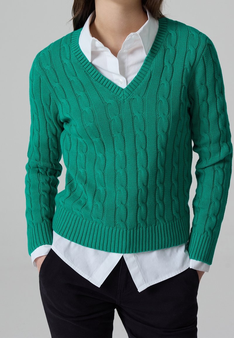 Personne portant un pull vert en maille torsadée à col en V par-dessus une chemise blanche boutonnée, avec un pantalon noir et les mains dans les poches.