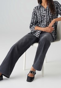 Femme assise sur une chaise blanche portant un chemisier à motifs noir et blanc, un jean noir délavé à jambes larges et des chaussures noires à boucle.