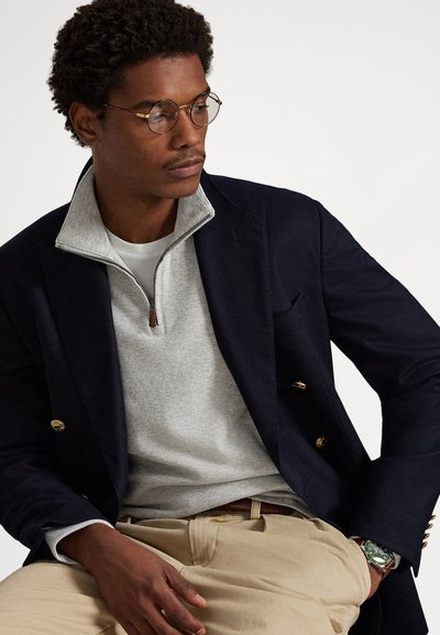 Mornarsko modro dvoredno srajčno jakno čez sivi pulover z zadrgo, kombinirano s kaki hlačami. Okrnjene s kroglastimi očali in uro za popoln videz.