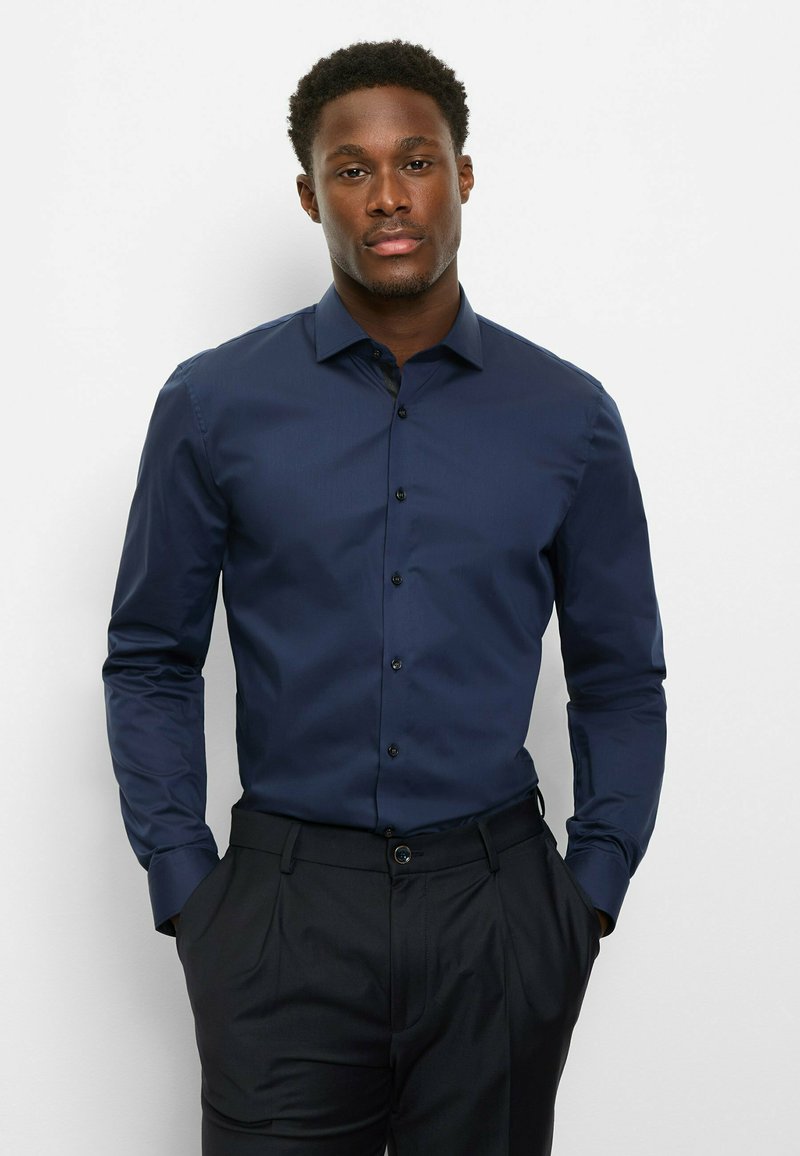 Chemise bleu marine à manches longues avec col boutonné, présentant une texture lisse et un léger éclat, associée à un pantalon foncé et ajusté.