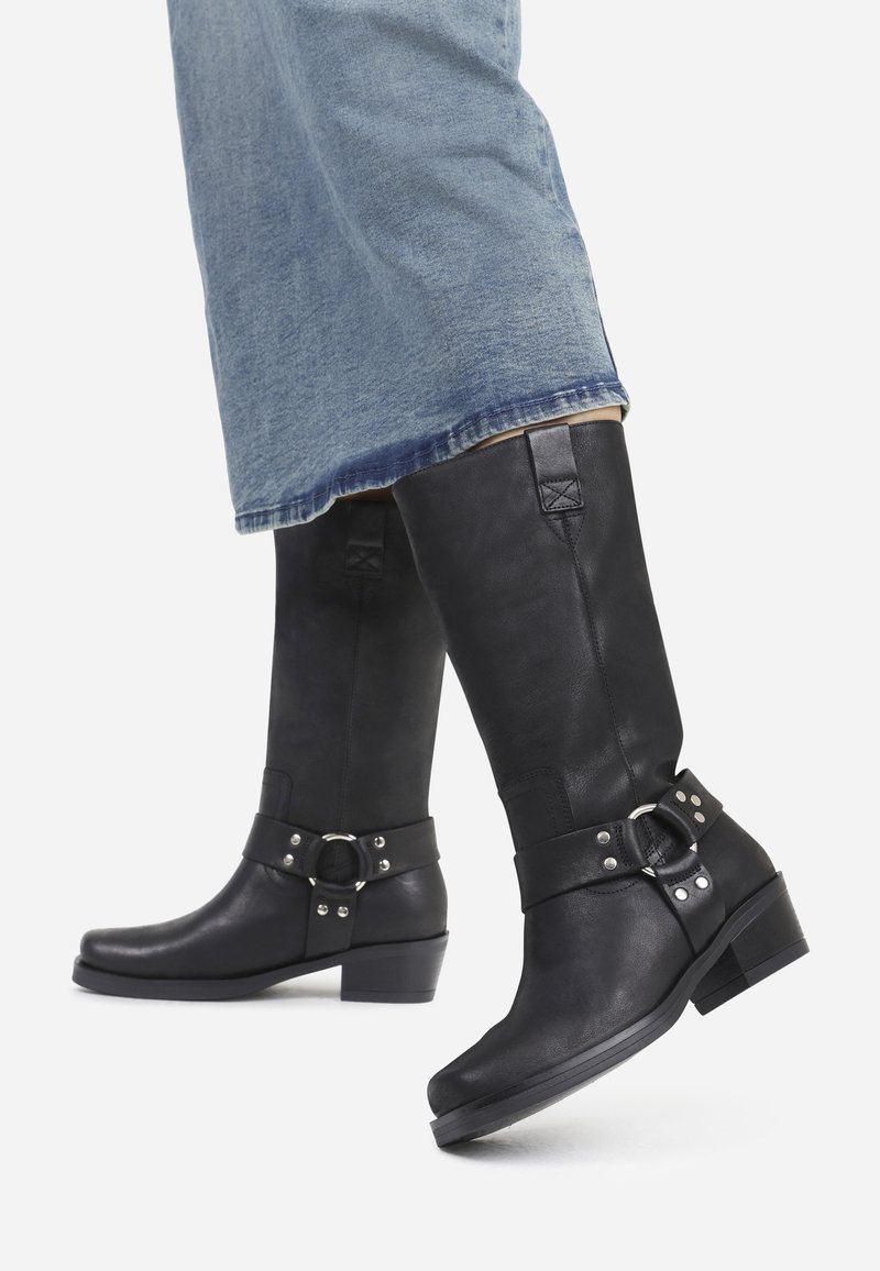 Botas altas de cuero negro hasta la rodilla con punta redonda, herrajes plateados, detalles de hebilla y un tacón bajo de bloque, combinadas con un denim de color azul claro.