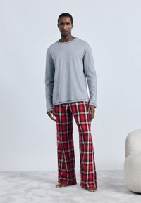 Grå långärmad skjorta i bomullsmaterial; röda och svarta rutiga pyjamasbyxor med lös passform; står på en ljus, heltäckningsmatta.