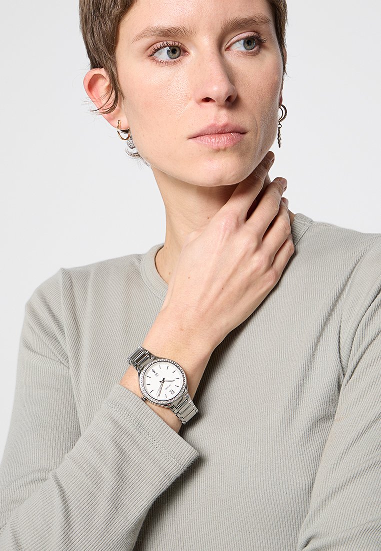 Montre en argent avec un cadran blanc rond, bracelet en métal argenté et repères horaires ; portée sur un poignet avec un haut à manches longues gris clair.