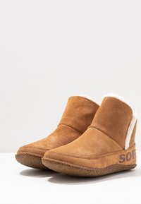 Tan-farbene Wildleder-Ankle Boots mit einem weichen weißen Fellfutter, runder Spitze und einer strukturierten Gummisohle, die an der Seite ein braunes Logo aufweist.