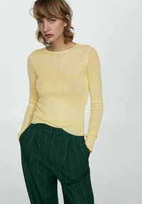 Rumena rebrasta majica z dolgimi rokavi in okroglo izrezom, v kombinaciji s temno zelenimi hlačami s črtami, ki imajo stranske žepe in sproščen kroj.