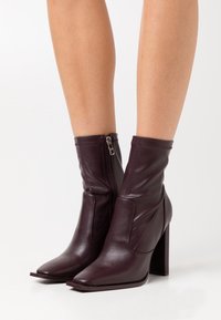 Botas de cuero de mujer en púrpura oscuro, con cremalleras laterales y tacones cuadrados, llevadas sobre piernas desnudas contra un fondo blanco.
