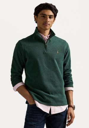 Green, ribbed quarter-zip sweater with a small logo, layered over a light pink striped shirt, paired with dark denim jeans.