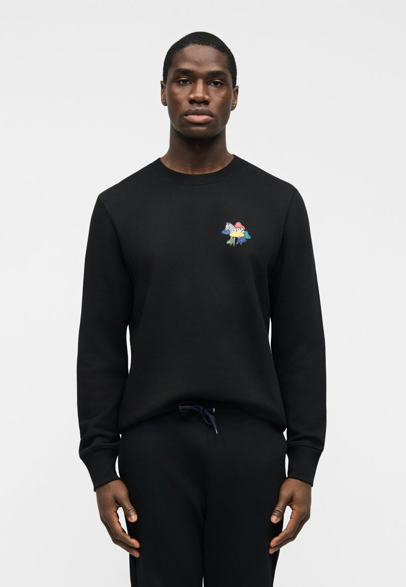 Άνδρας που φοράει μαύρο μακρυμάνικο πουκάμισο και παντελόνι με πολύχρωμο σχέδιο ζέβρας και μανιταριών στο στήθος, στέκεται μπροστά σε απλό ανοιχτό φόντο.