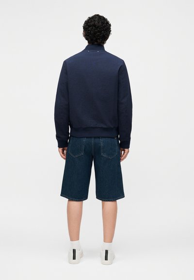 Homme aux cheveux noirs bouclés, debout de dos, portant une veste bleu marine, un short en jean jusqu'aux coudes, des chaussettes blanches et des baskets blanches avec des bandes noires.