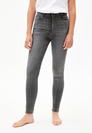 Persona con pantalones vaqueros pitillo negros desgastados y una camiseta blanca, de pie descalza sobre una superficie blanca.